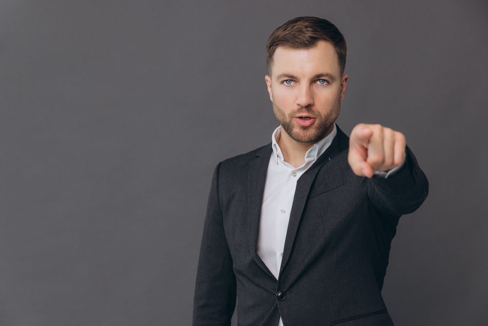 Confident businessman in a suit pointing directly at the you against a plain grey background.