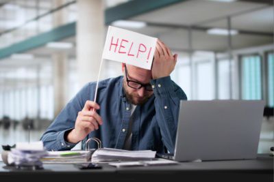 Overworked professional holding help sign at cluttered desk representing businesses without internal HR recruiting expertise
